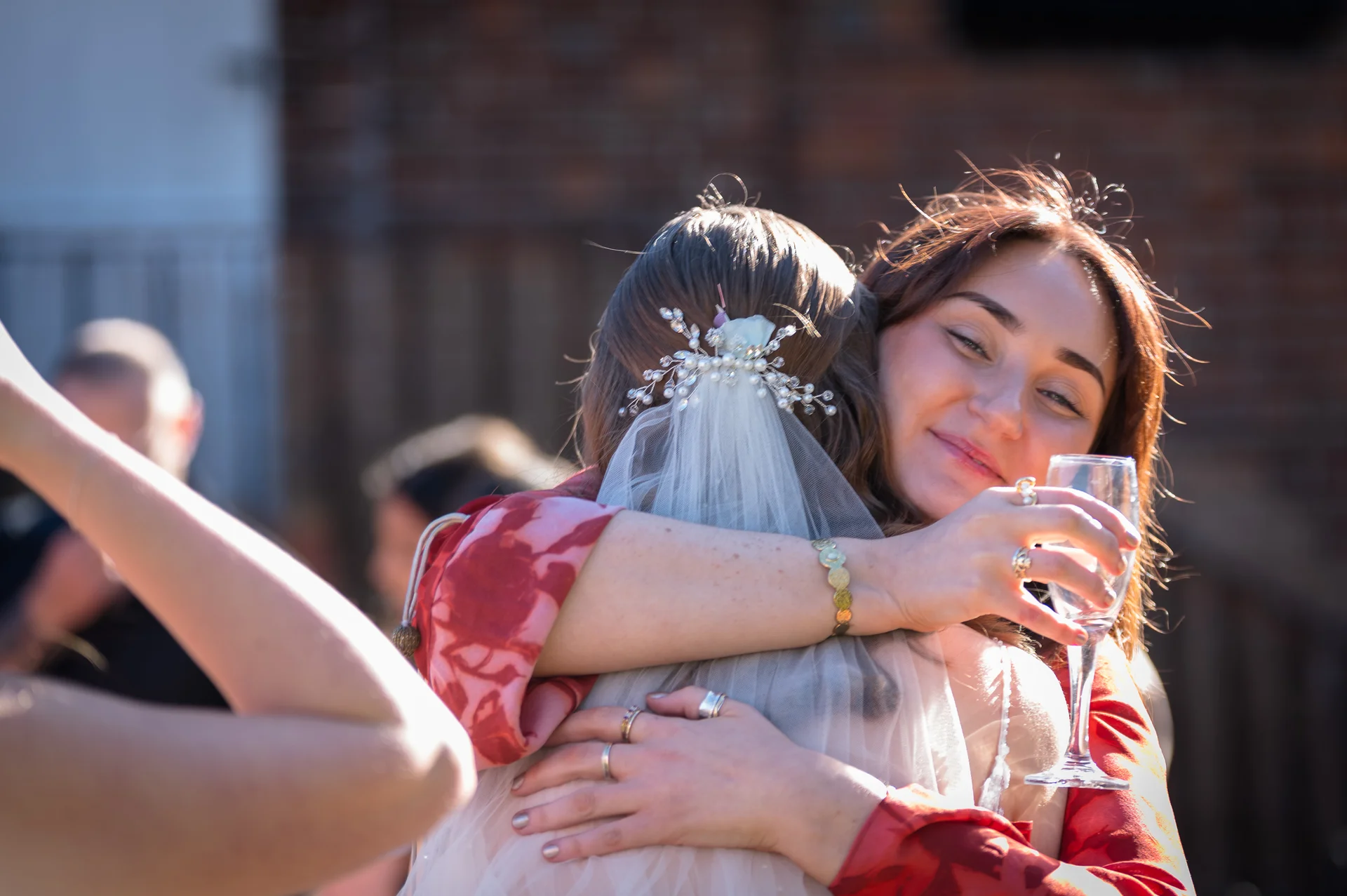 A smiling guest embraces a bride wearing a delicate veil and hair comb while holding a glass of champagne during a sunny drinks reception.