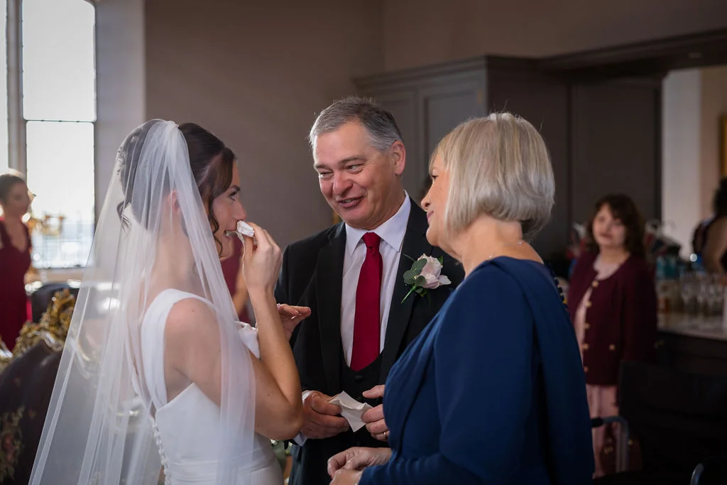 An emotional bride wipes a tear away while sharing a tender moment with her smiling father and mother before the ceremony.