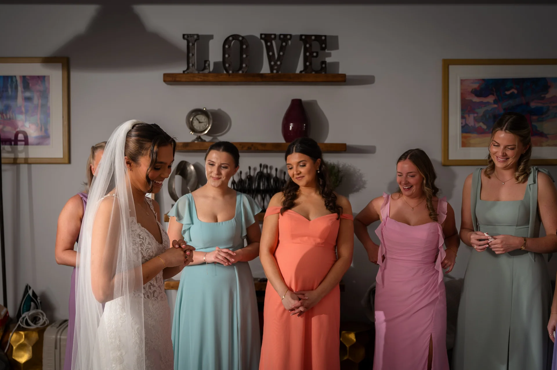 A smiling bride wearing a lace gown and veil looks down at her hands, surrounded by her bridesmaids in colourful pastel dresses.