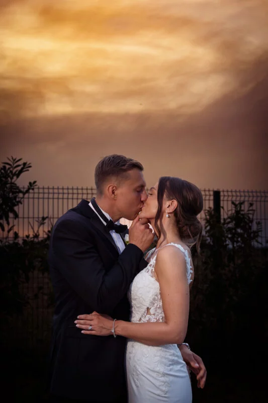 A bride and groom kissing in front of a fence at sunset.