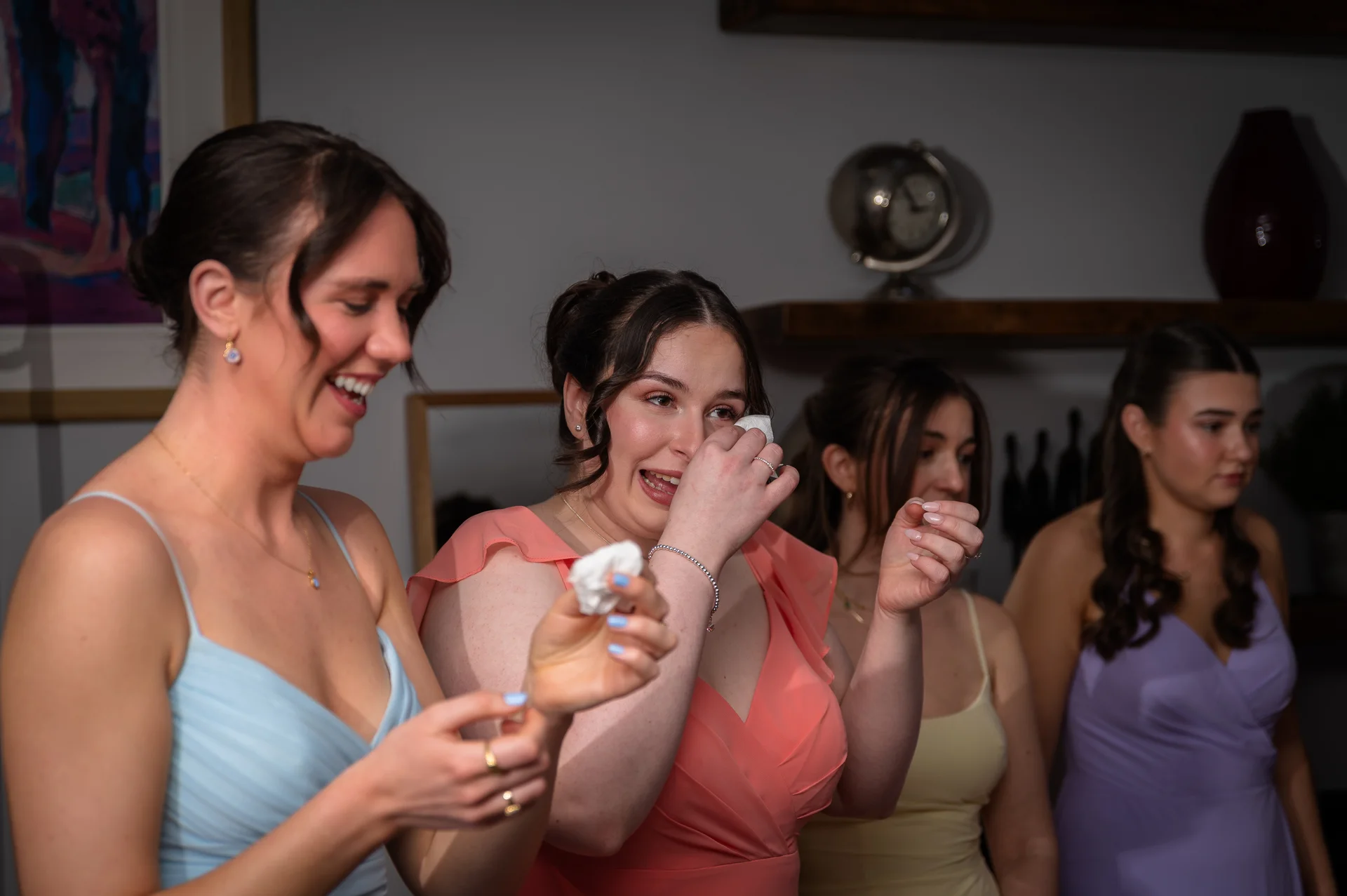A group of bridesmaids in colourful dresses share an emotional and joyful moment, wiping happy tears while laughing together indoors.