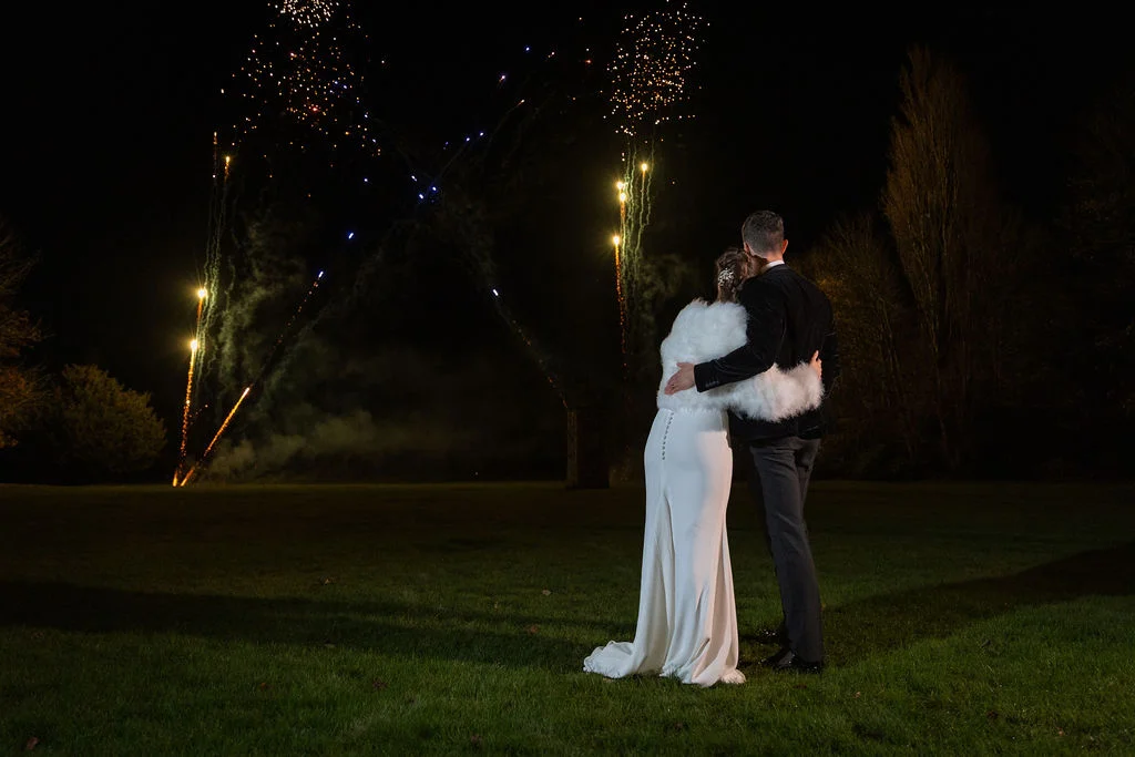 A newlywed couple embraces on a lawn at night, watching a vibrant fireworks display light up the dark sky.