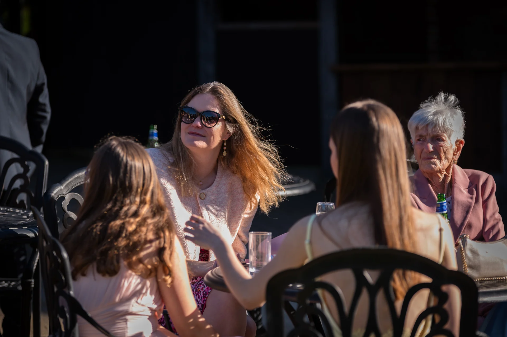 Four female wedding guests spanning multiple generations sit together outdoors at black metal tables, enjoying the sunny afternoon reception.