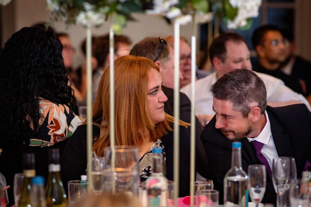 Wedding guests converse and enjoy a meal at tables decorated with tall floral centrepieces and glassware during the wedding breakfast.