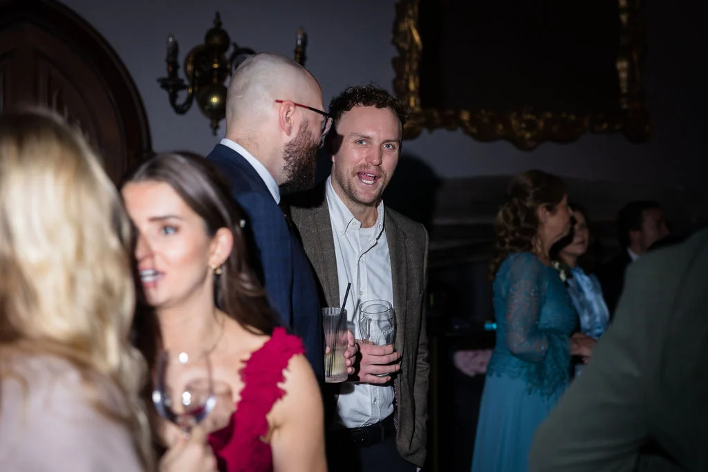 Wedding guests mingle and chat during an evening drinks reception inside a dimly lit historic venue, holding glasses and laughing.