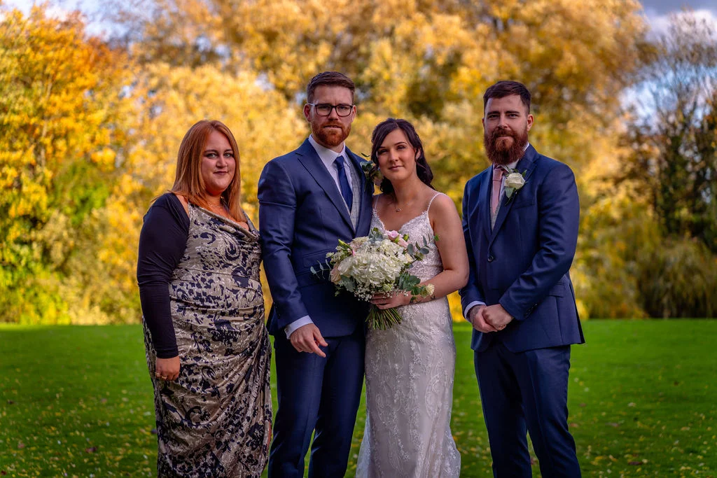 A bridal party comprising a bride holding a white bouquet, a groom, another man in a navy suit, and a woman in a patterned dress stand together outdoors against a backdrop of vibrant autumn foliage.