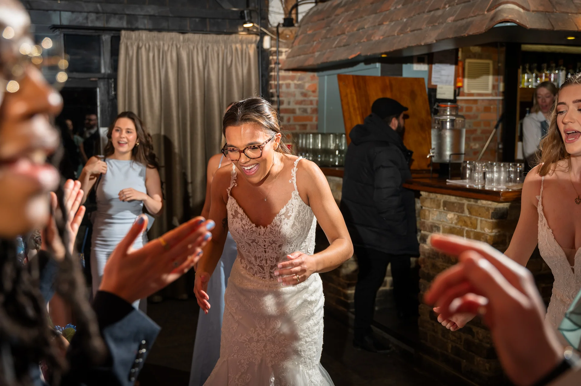 A joyful bride wearing glasses and a lace fit and flare gown laughs excitedly on the dance floor surrounded by cheering guests in a rustic pub venue.