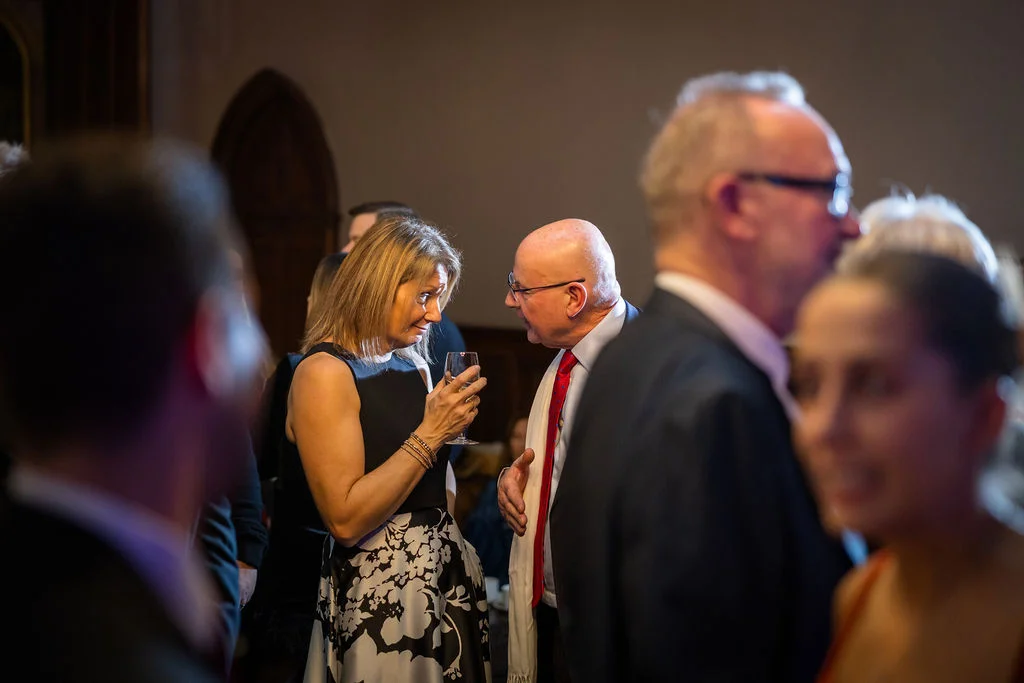 Wedding guests mingle and converse in a softly lit interior space, captured candidly among a gathering of family and friends.