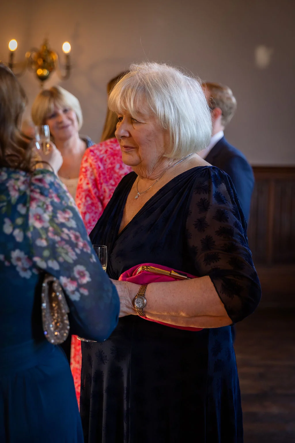 An elegant older woman with short white hair wearing a dark patterned dress holds a pink clutch bag while mingling during the wedding reception.