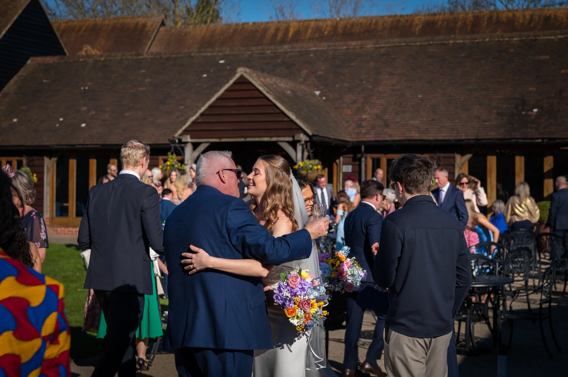 A bride embraces a guest wearing a blue suit outside a rustic barn venue during a sunlit drinks reception.