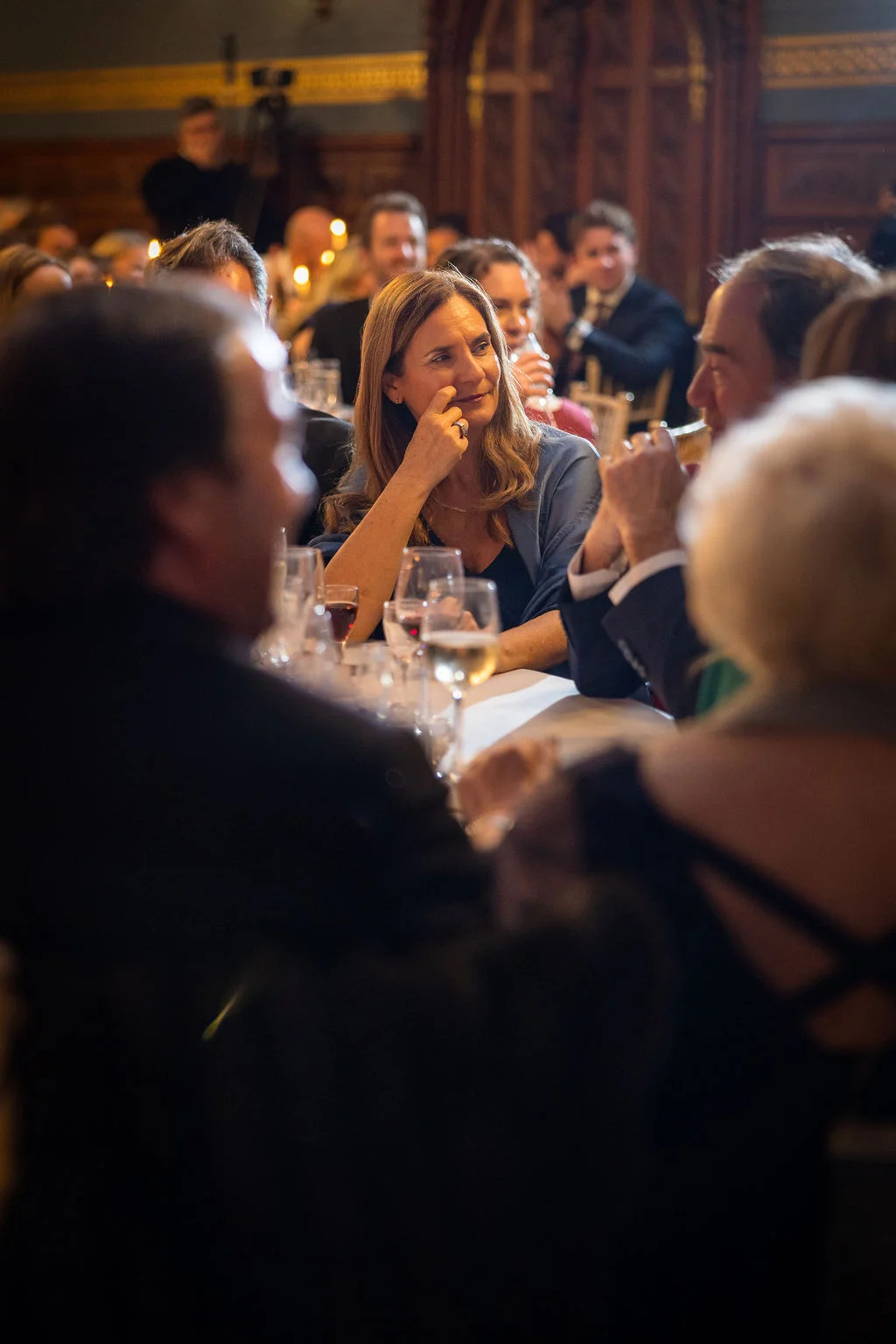 Guests are captured in a candid moment during wedding speeches, with a woman resting her chin on her hand and smiling while seated at a banquet table.
