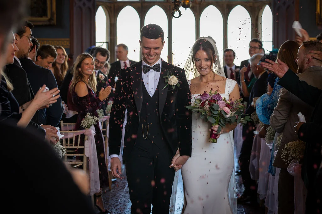 A newlywed couple walks down the aisle hand in hand while smiling guests shower them with a cascade of red confetti inside a historic venue with tall arched windows.