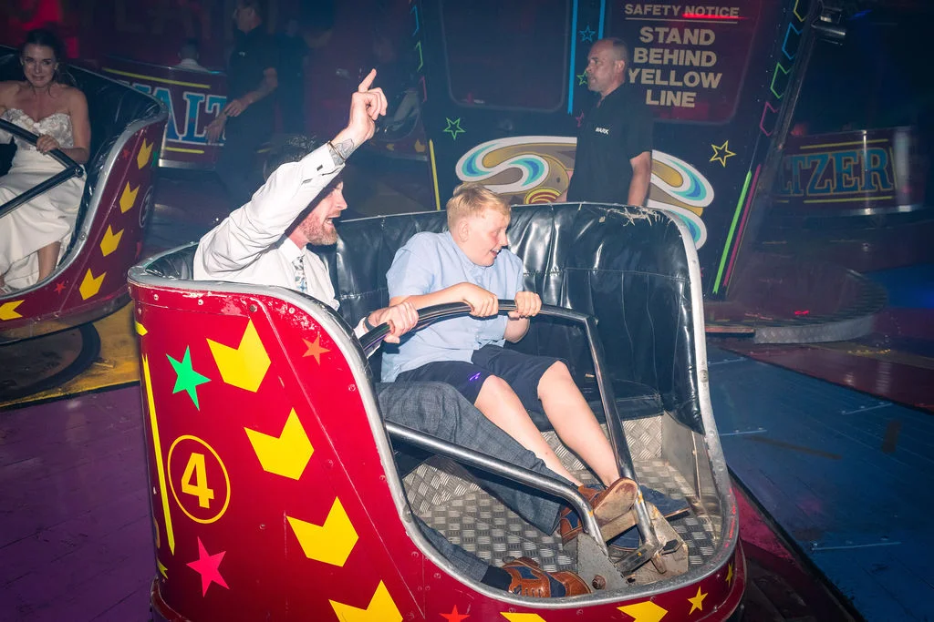 A groom and a young guest having fun riding a fairground car at an evening wedding reception with colorful lights.