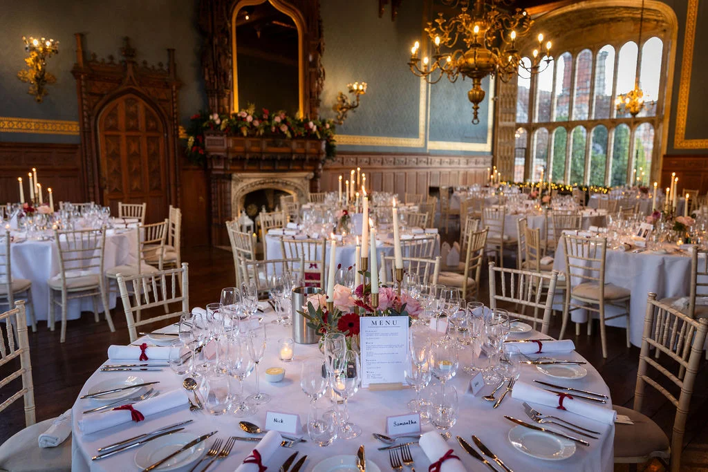 An elegantly set round wedding table sits inside a grand historic hall featuring tall arched windows, a large ornate chandelier, and rich wood panelling.