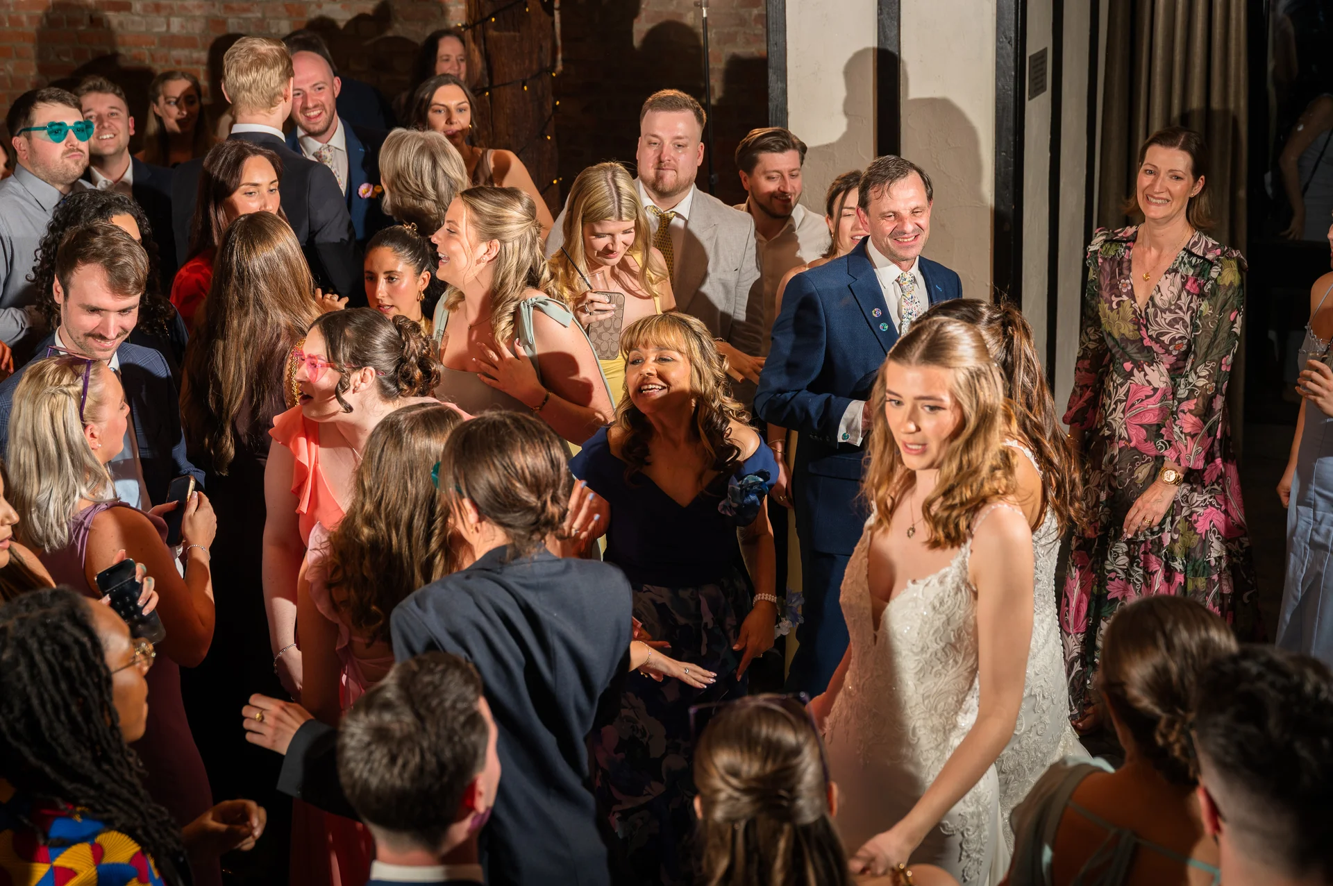 A bride in a white gown and her smiling guests enjoy the bustling evening wedding reception amidst a crowd of cheering attendees.