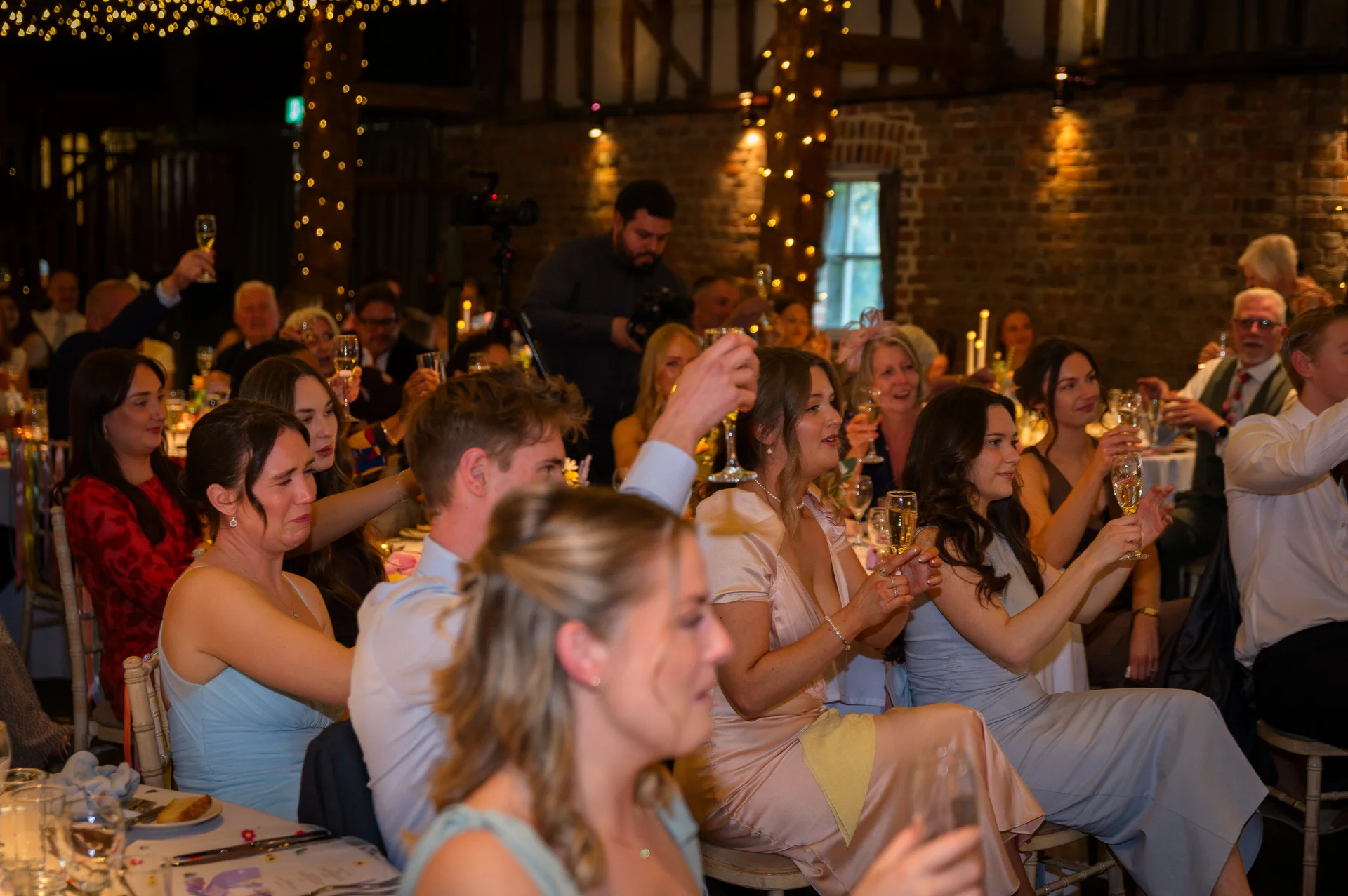 Wedding guests raise champagne glasses in toast during speeches inside a rustic barn venue illuminated by fairy lights.
