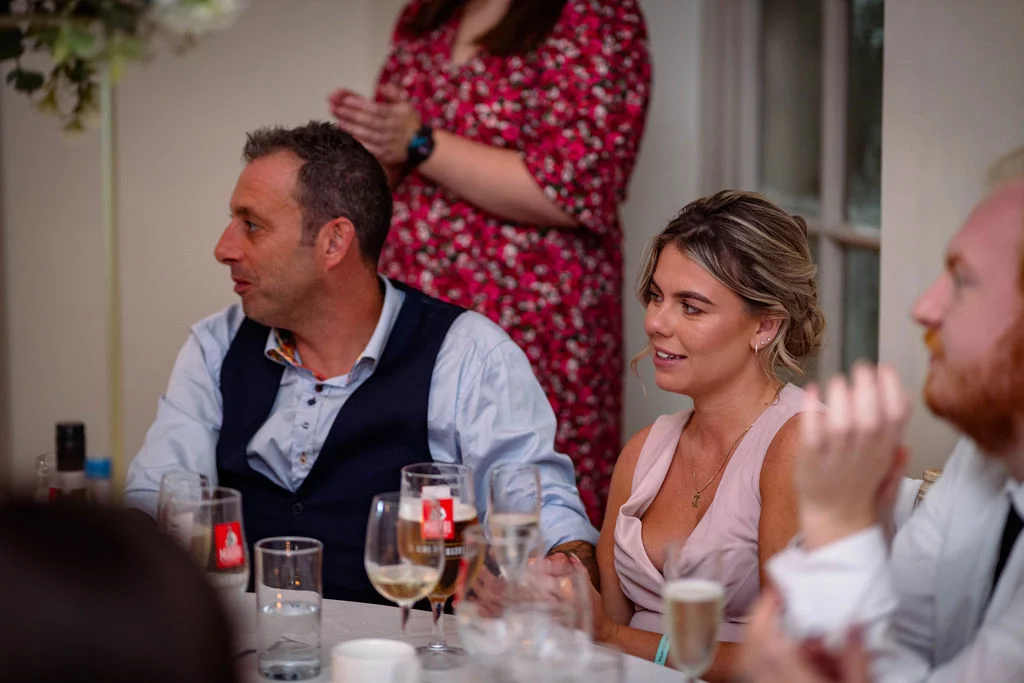 Wedding guests smiling and listening intently during speeches at a wedding breakfast reception table.