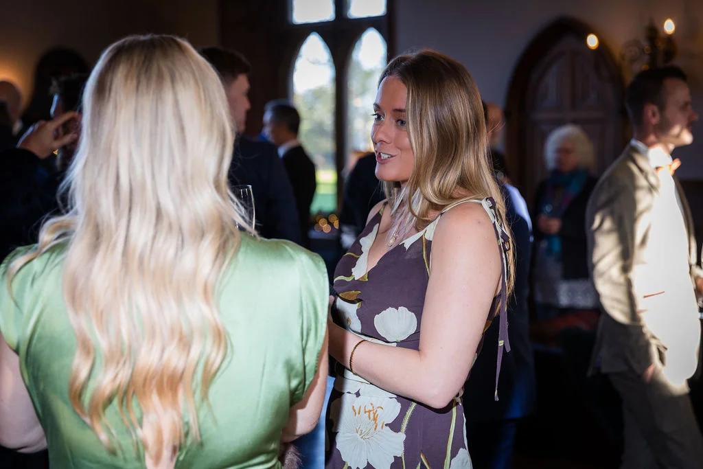 Wedding guests mingling and chatting together during a drinks reception inside a historic venue illuminated by soft window light.