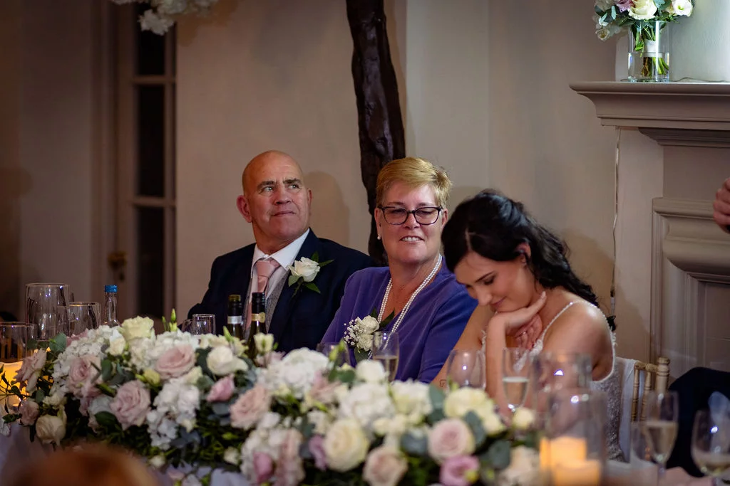 A bride and her parents are seated at the top table during wedding speeches, with the bride smiling while looking down behind a lush floral centrepiece.