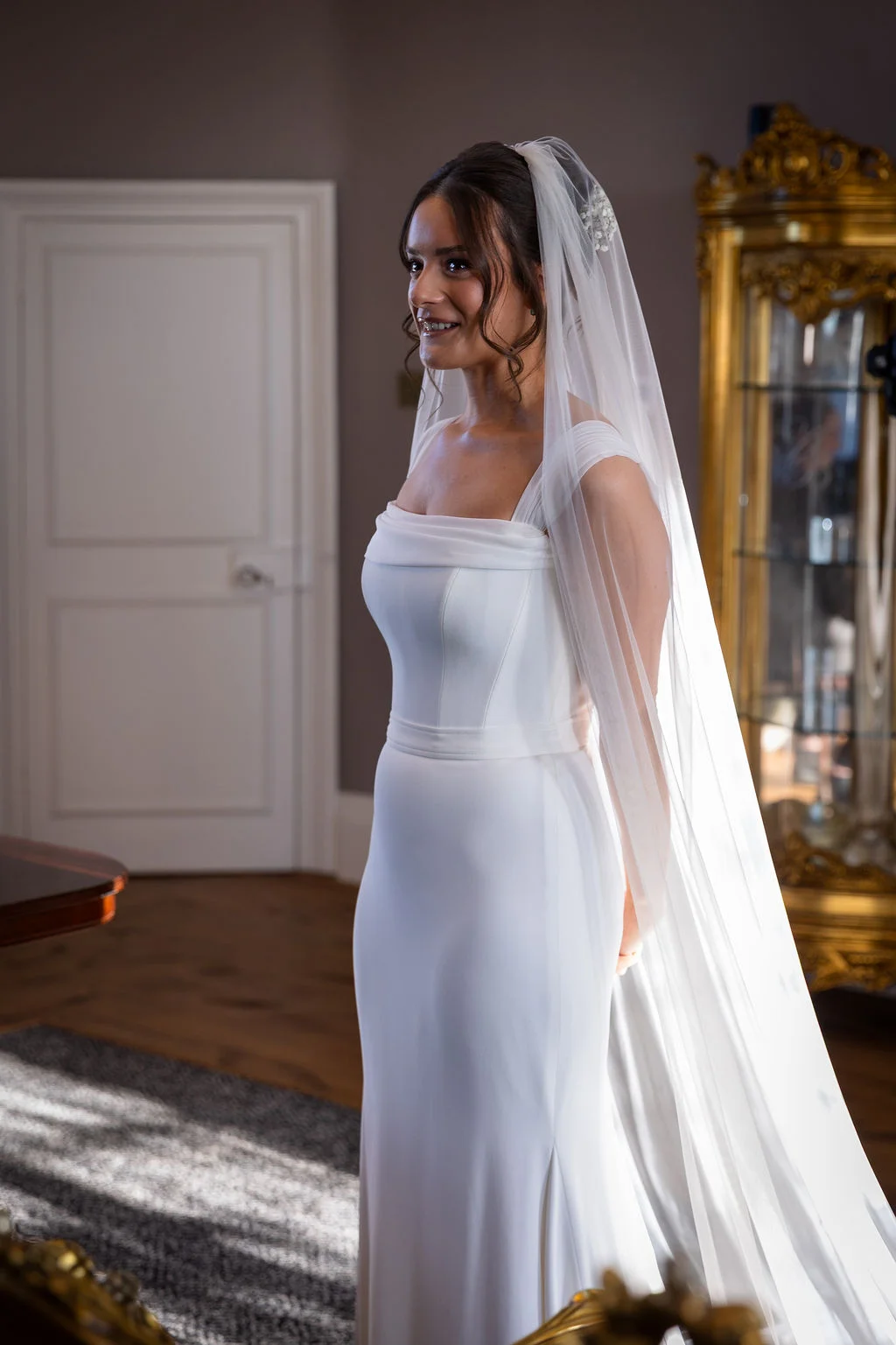 A smiling bride wearing a structured white crepe gown with a square neckline and a long veil stands indoors illuminated by soft natural light.