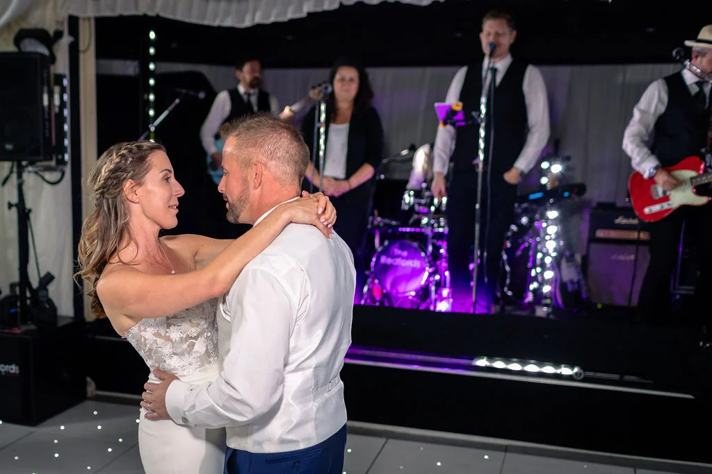 A bride and groom share a tender moment dancing on an illuminated dance floor while a live band performs on stage in the background.