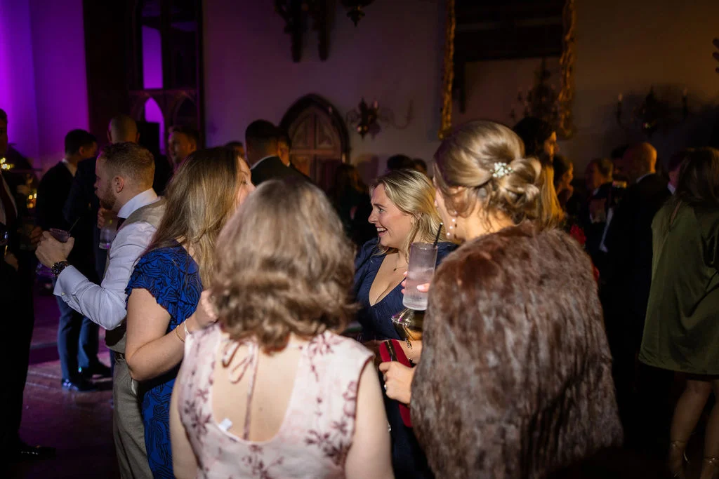 Wedding guests mingle and enjoy drinks during an evening reception illuminated by moody coloured lighting.