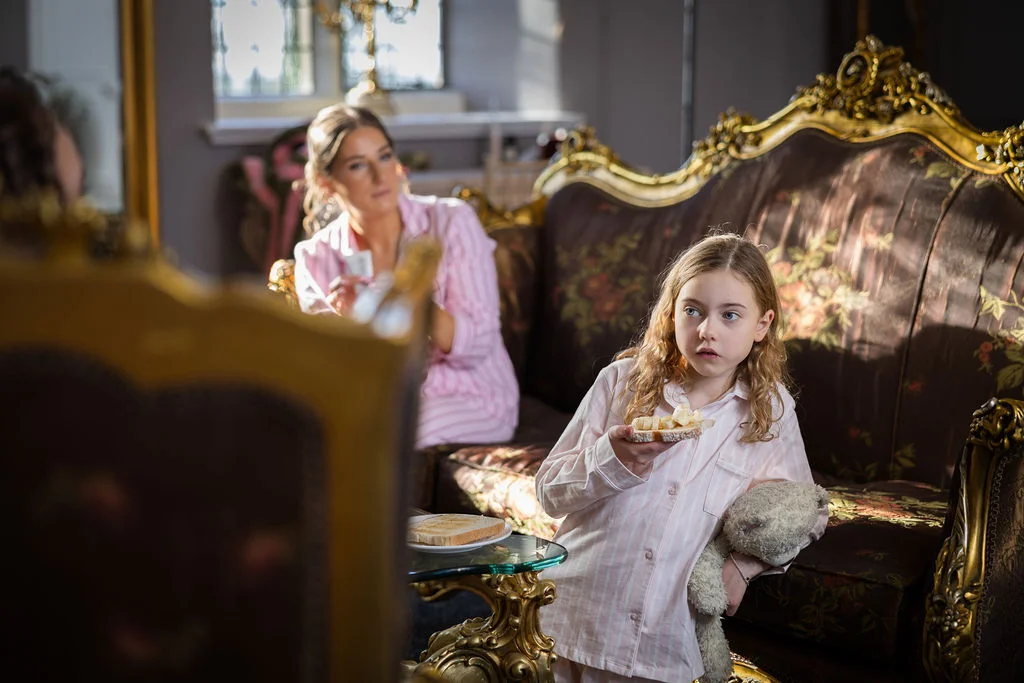 A young flower girl in striped pyjamas holds a piece of toast and a stuffed toy while getting ready in a grand room with an ornate sofa.