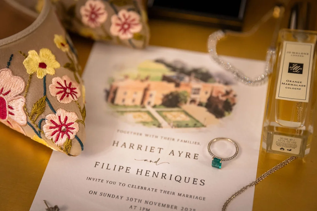 An elegant bridal flat lay featuring an illustrated wedding invitation, embroidered floral shoes, a glittering engagement ring with a green gemstone, and a bottle of cologne.
