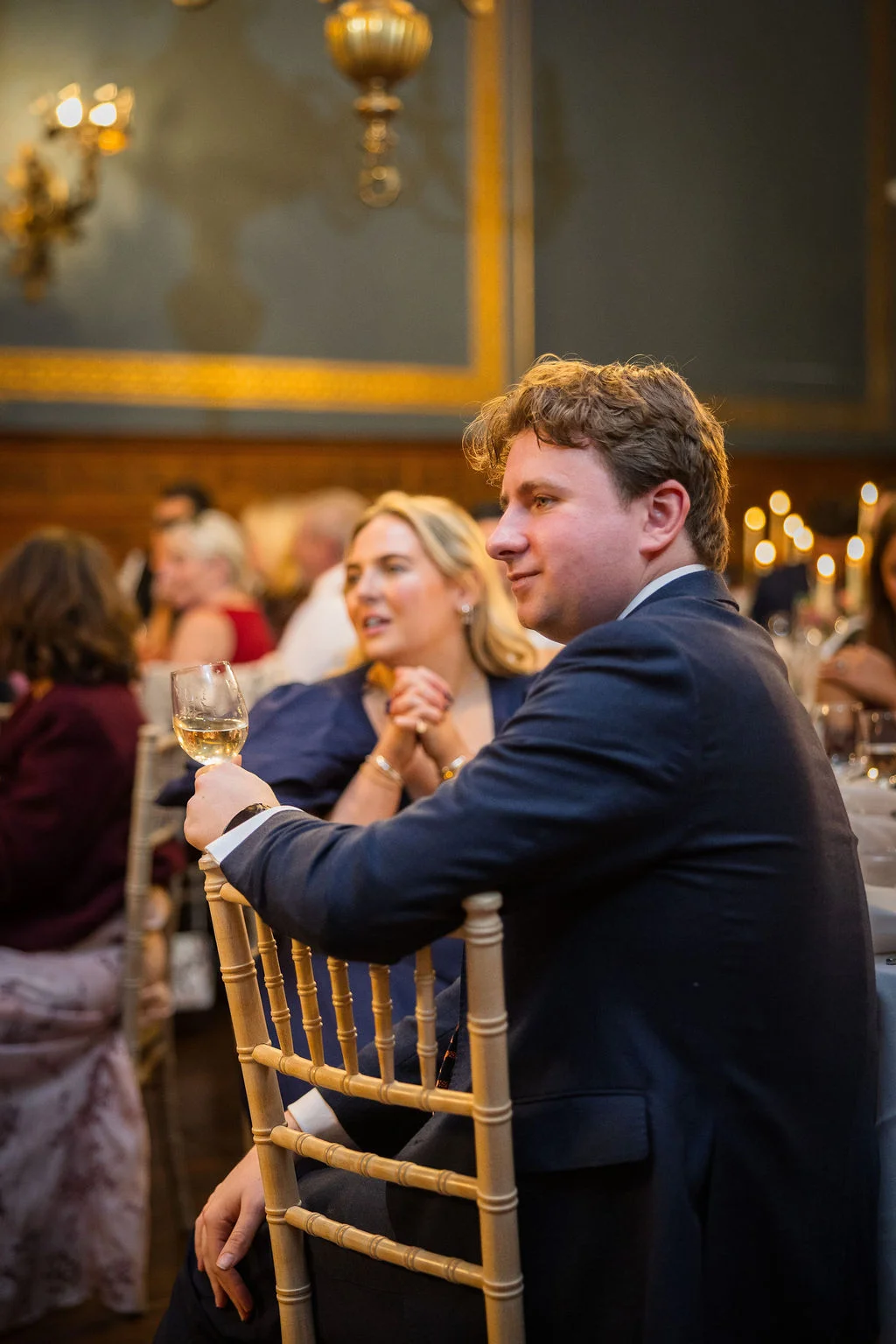A guest in a navy suit holds a glass of wine while listening attentively during wedding speeches in an elegant wood-panelled room.