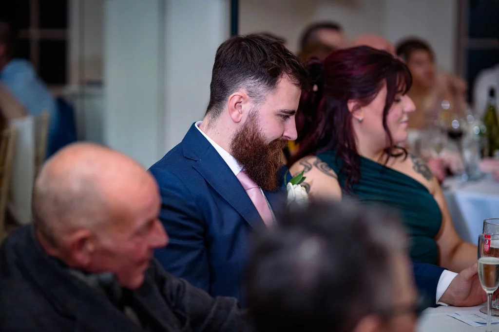 A bearded groom in a navy suit and pink tie sits attentively during the wedding breakfast alongside guests at a reception table.