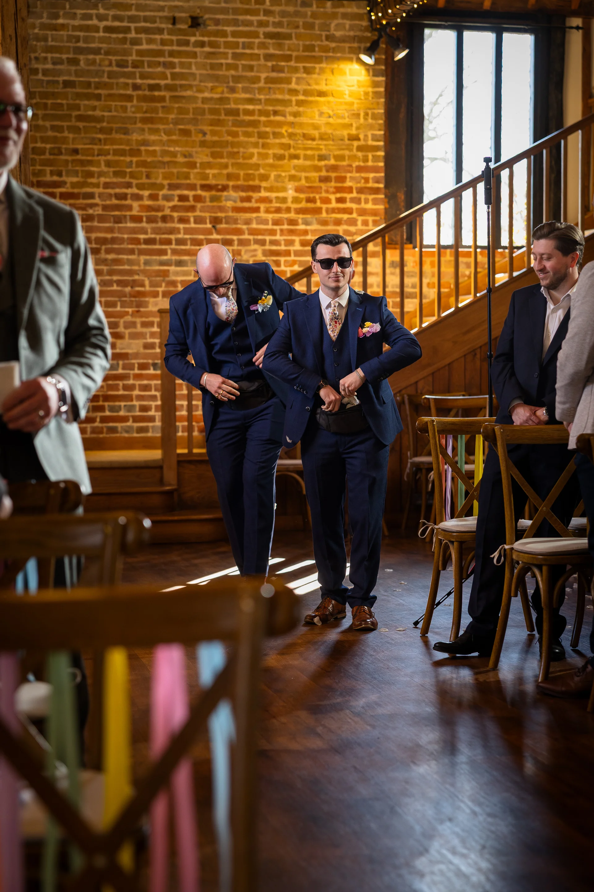 A group of groomsmen wearing navy suits and patterned ties share a laugh inside a rustic barn venue with exposed brick and wooden details.