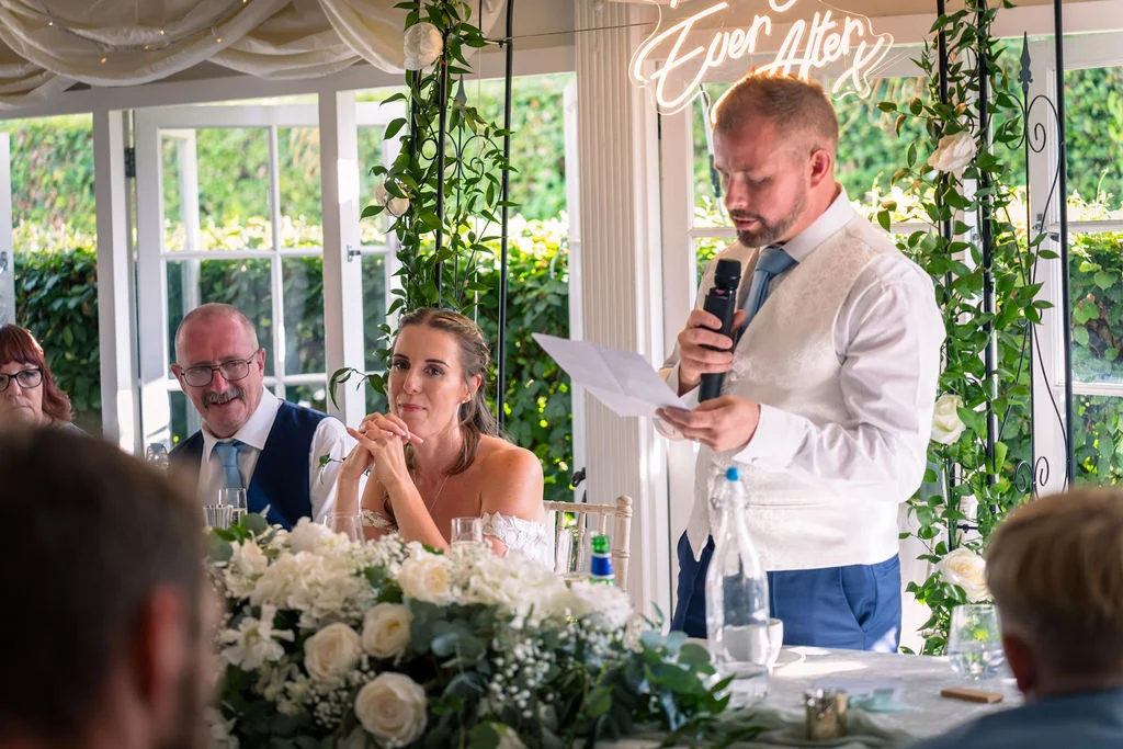 A groom stands at the top table delivering a wedding speech while holding notes and a microphone, with the bride and guests seated nearby against a backdrop of white floral arrangements and a neon sign.