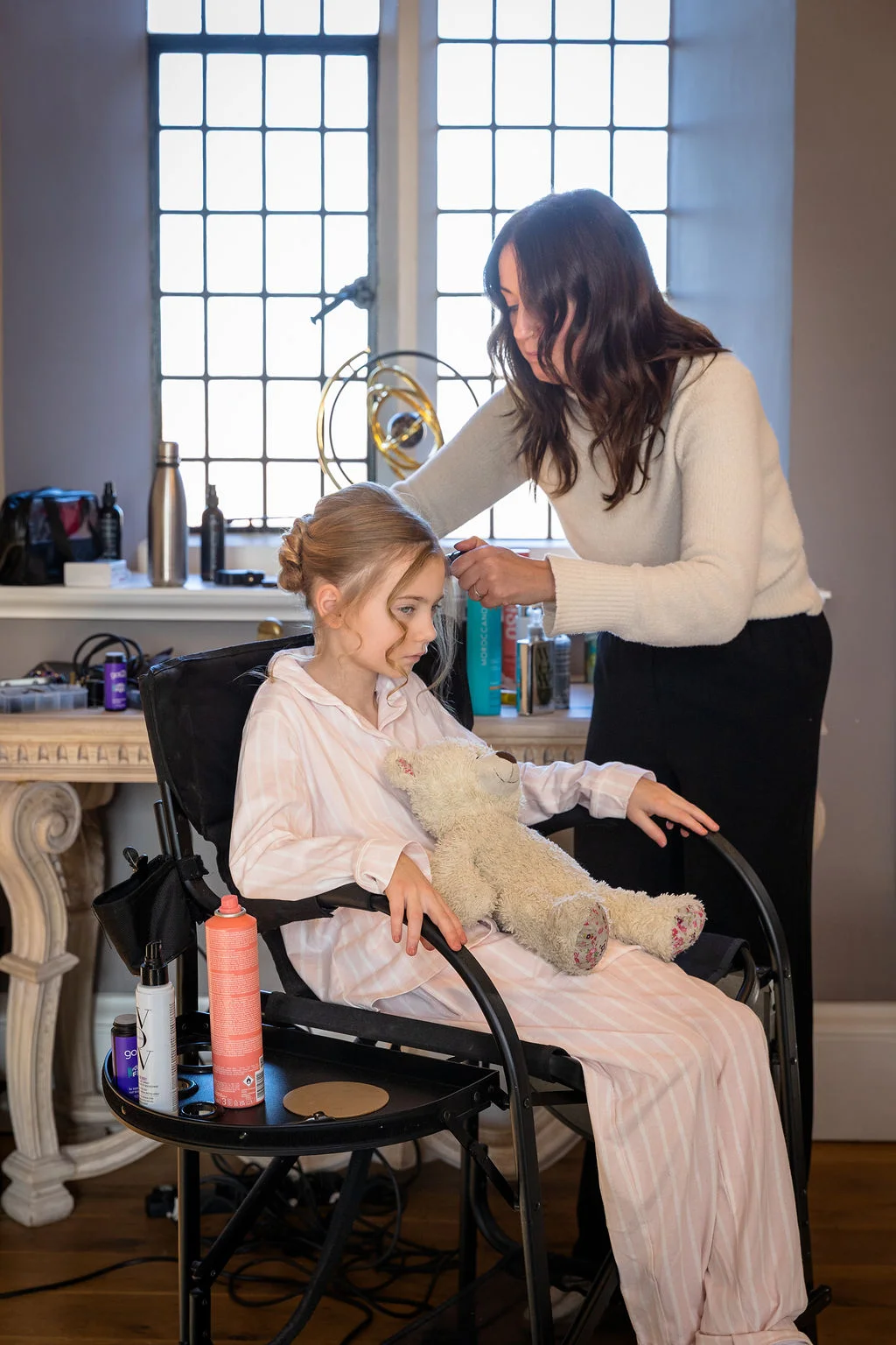 A young bridesmaid sits in a makeup chair wearing striped pyjamas while a stylist works on her hair near a bright window.