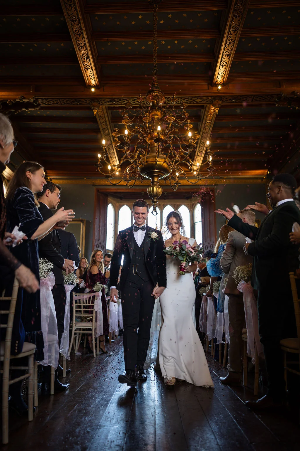 Newlyweds walk down the aisle hand in hand beneath a grand chandelier as guests shower them with confetti in a historic hall.