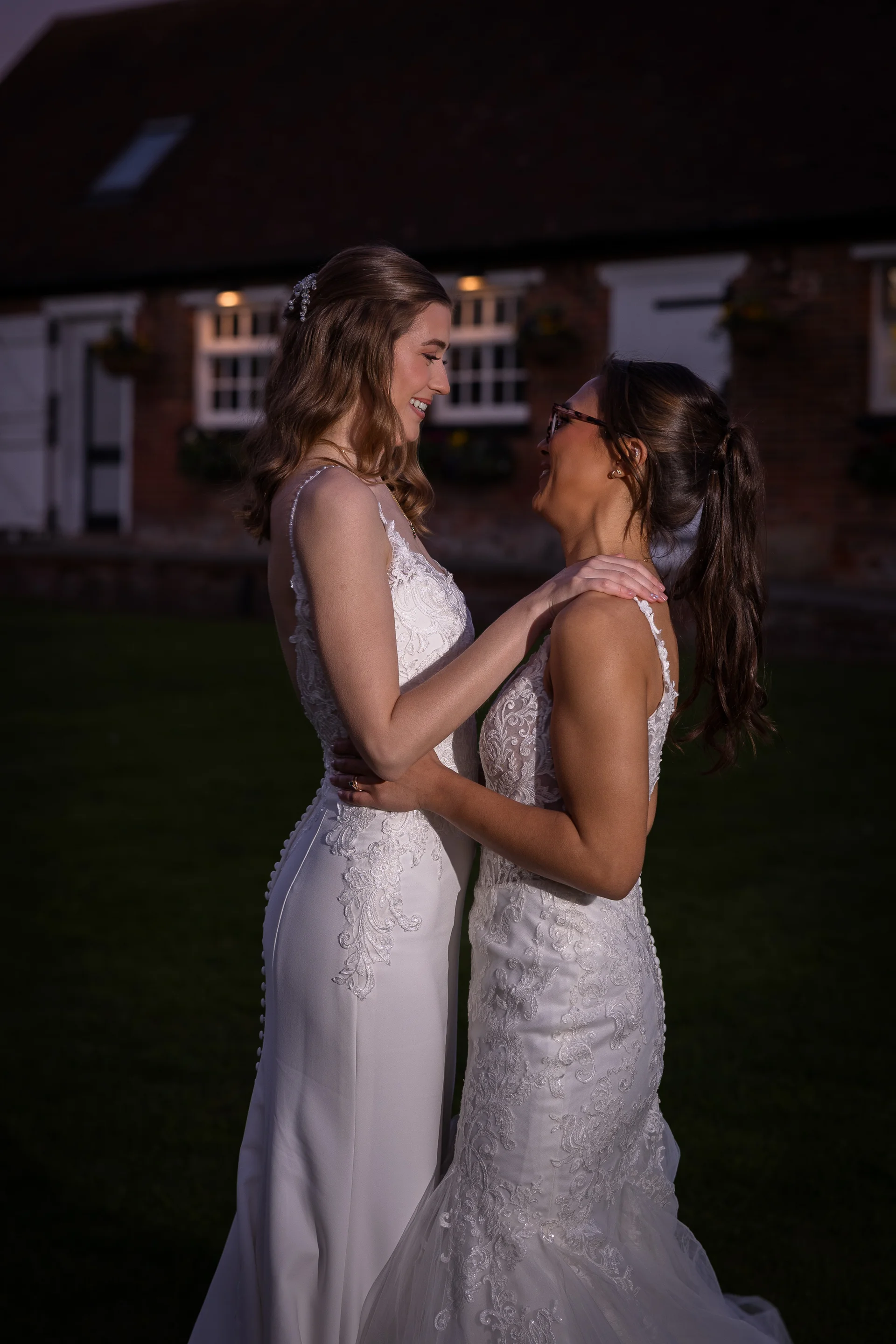 Two smiling brides in matching lace wedding dresses embrace tenderly outside a rustic barn at dusk.