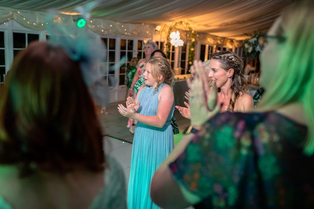 Wedding guests clap and cheer enthusiastically inside a marquee illuminated by soft lighting during the evening reception.