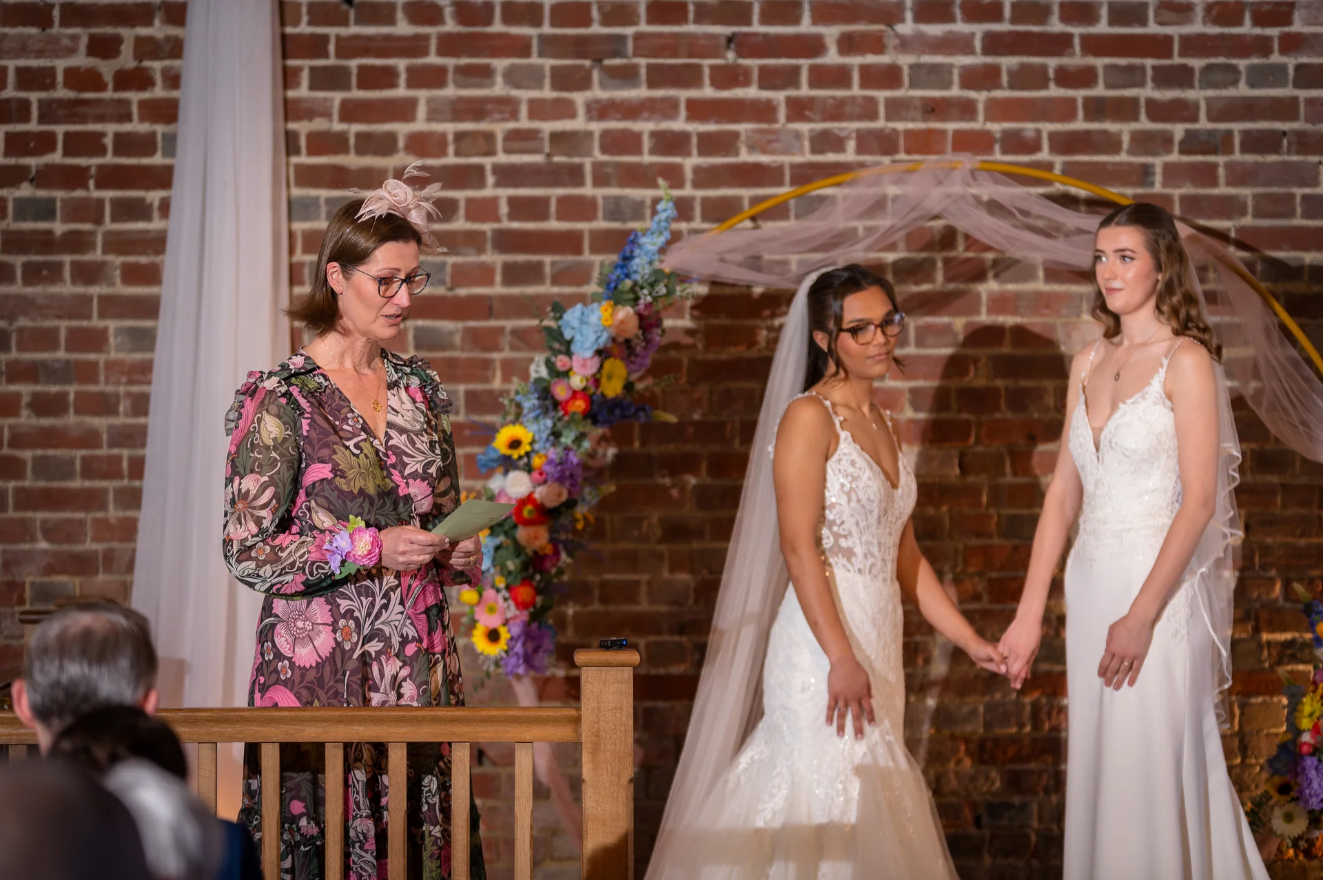 A celebrant reads from a card during a ceremony in front of an exposed brick wall, as two brides hold hands beneath a floral arch.