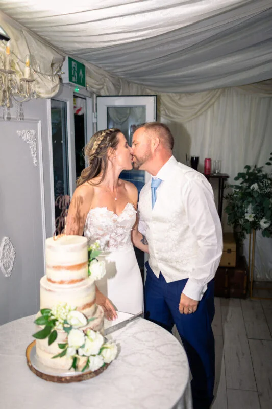 A newlywed couple share a tender kiss over a semi-naked three-tiered wedding cake adorned with white florals inside a draped marquee.