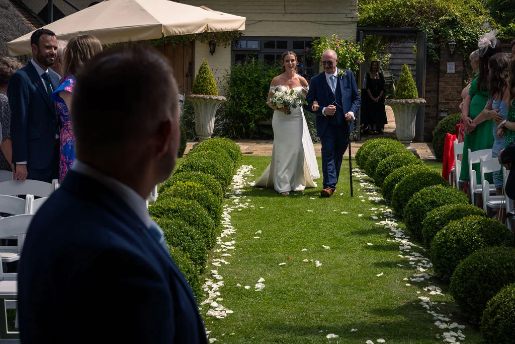 An over the shoulder shot of a groom watching his bride walk down a garden aisle lined with topiary bushes and white petals, escorted by her father.