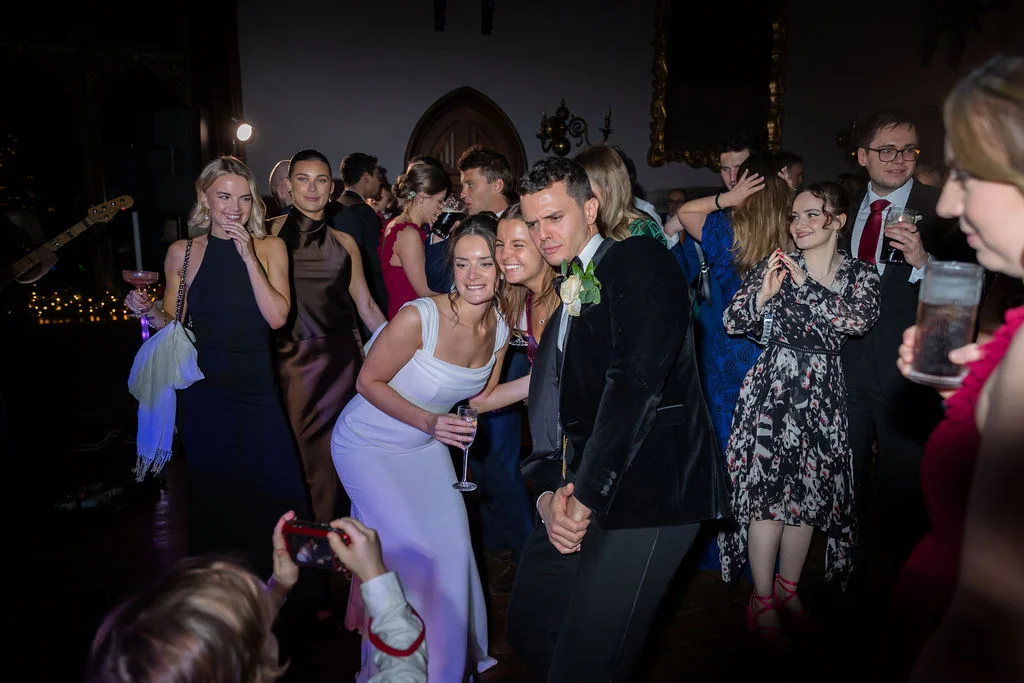 The bride and groom share a joyful dance surrounded by laughing wedding guests in a dimly lit evening reception venue.