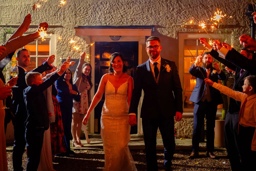 A smiling bride and groom walk hand in hand through a tunnel of glowing sparklers held by cheering guests at night.