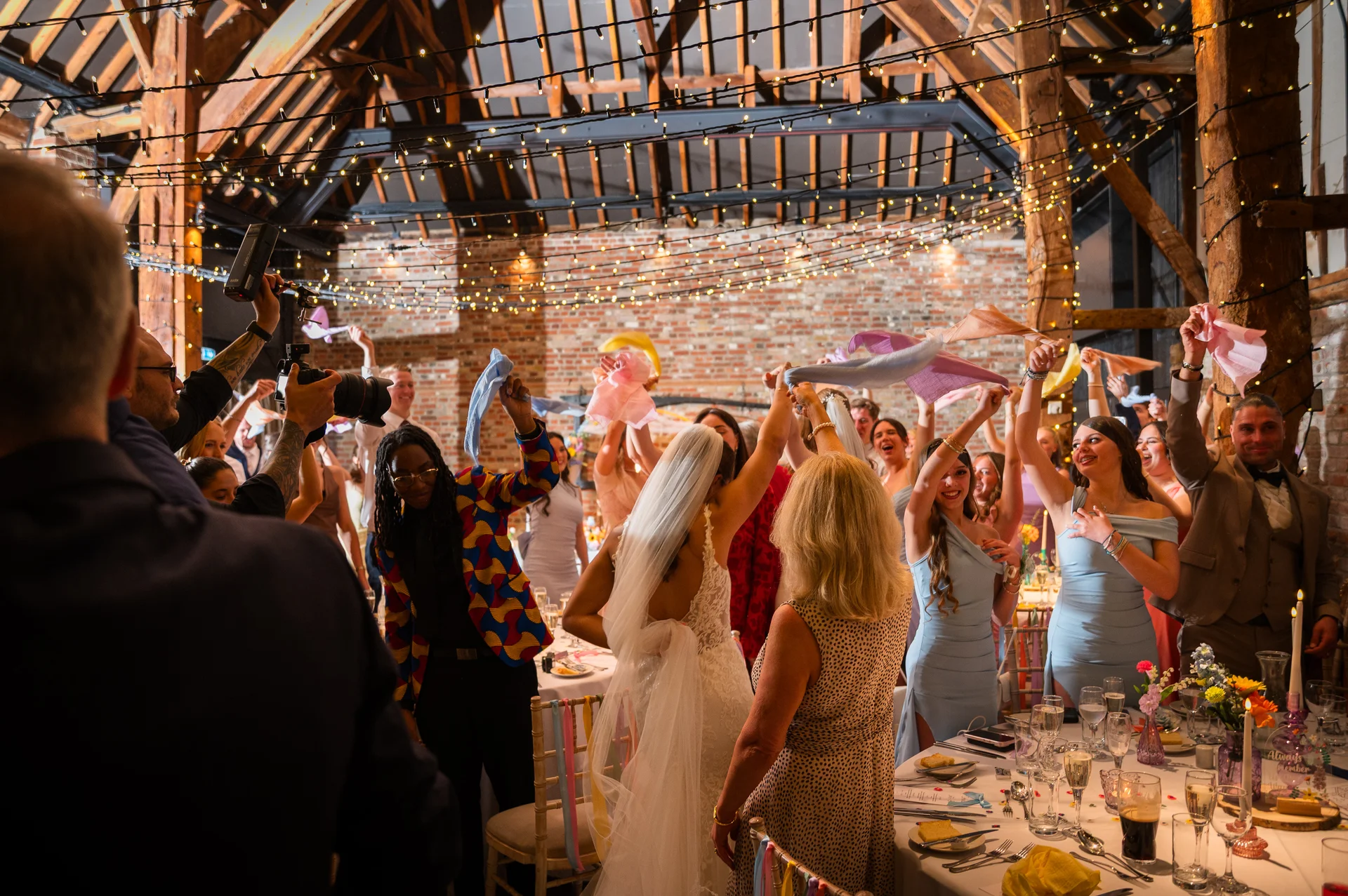 A joyous wedding celebration takes place inside a rustic barn illuminated by fairy lights, where guests wave napkins in the air around the newlyweds.