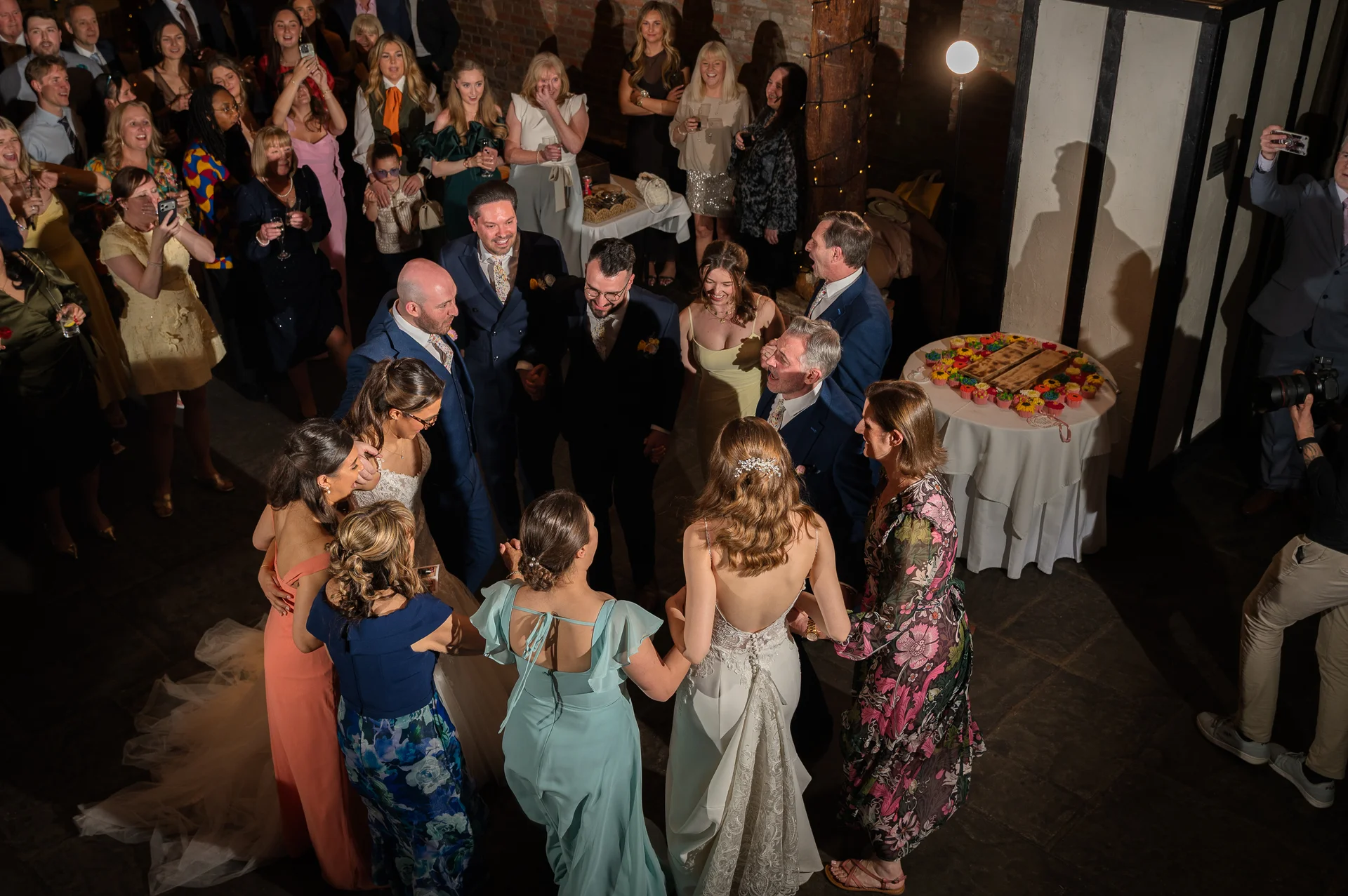 A high-angle candid shot of a lively wedding party and guests dancing and laughing together in a brightly lit indoor venue.