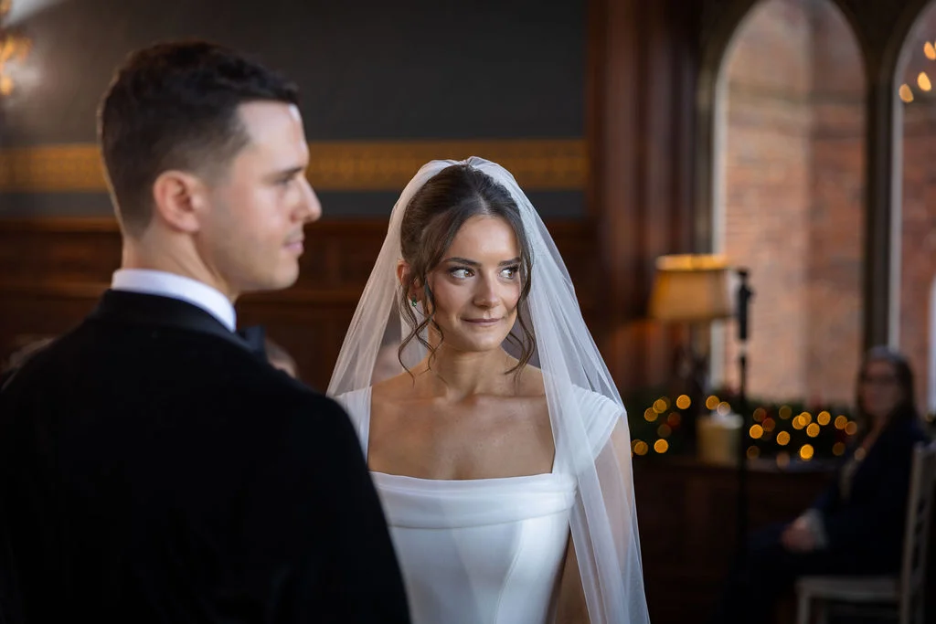 A newly married couple shares a tender glance during their indoor wedding ceremony, with the bride wearing a sleek gown and veil.