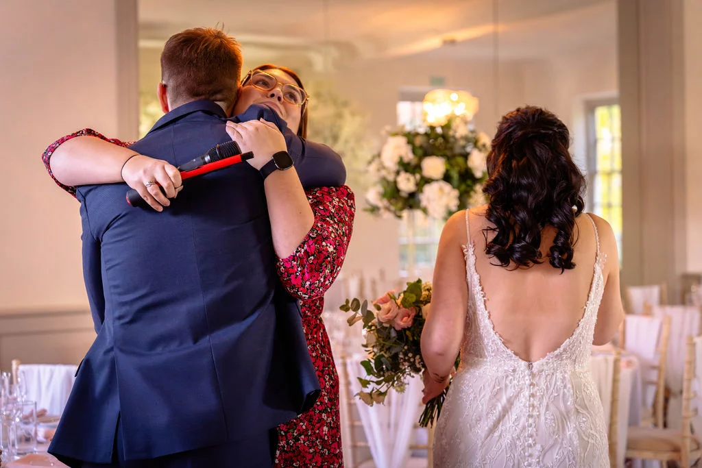 A guest in a red floral dress hugs someone holding a microphone while a bride in a backless lace gown watches in the mirror reflection.