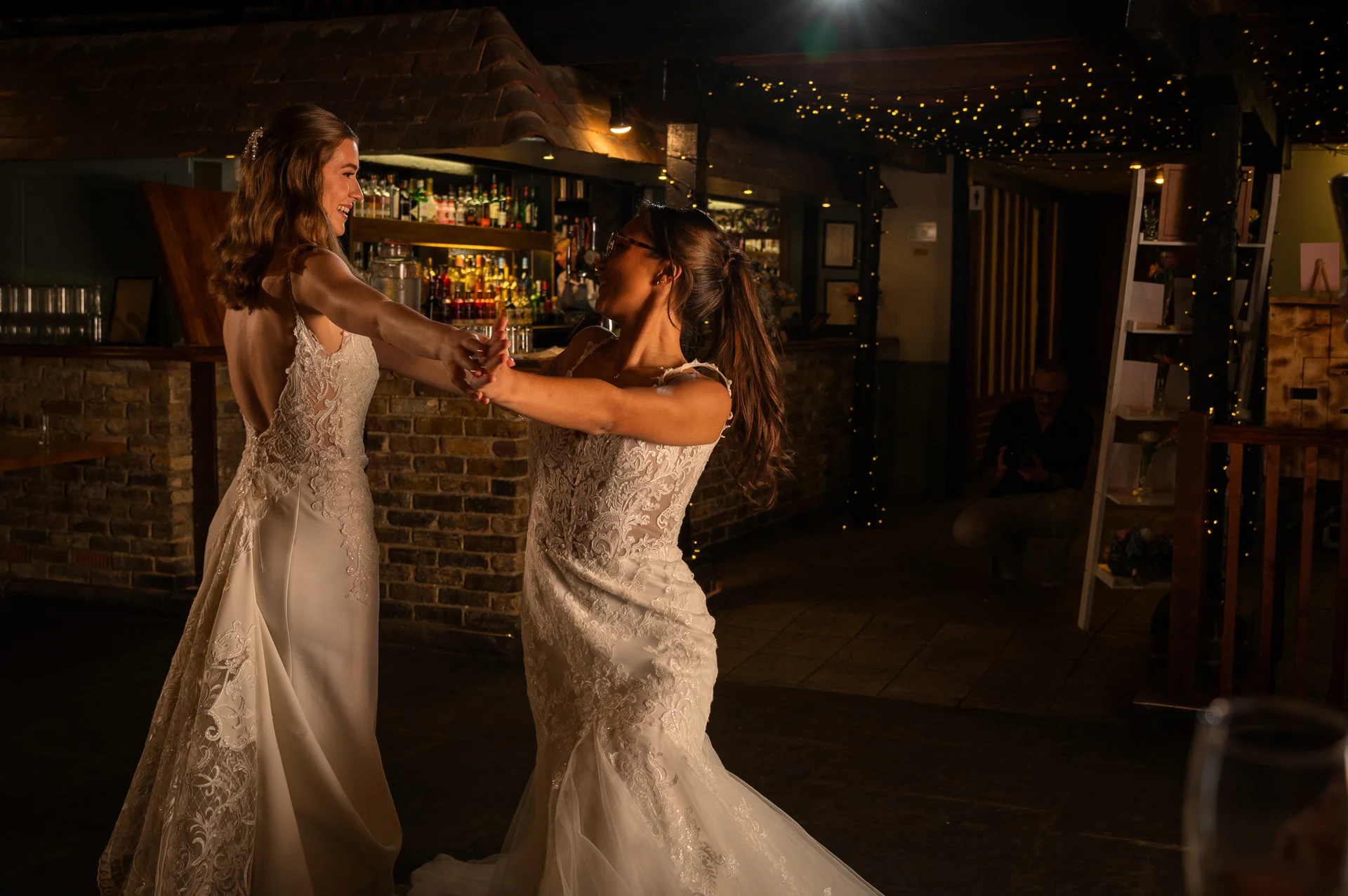 Two brides share a joyful first dance inside a cosy pub venue illuminated by fairy lights, wearing stunning lace wedding gowns.