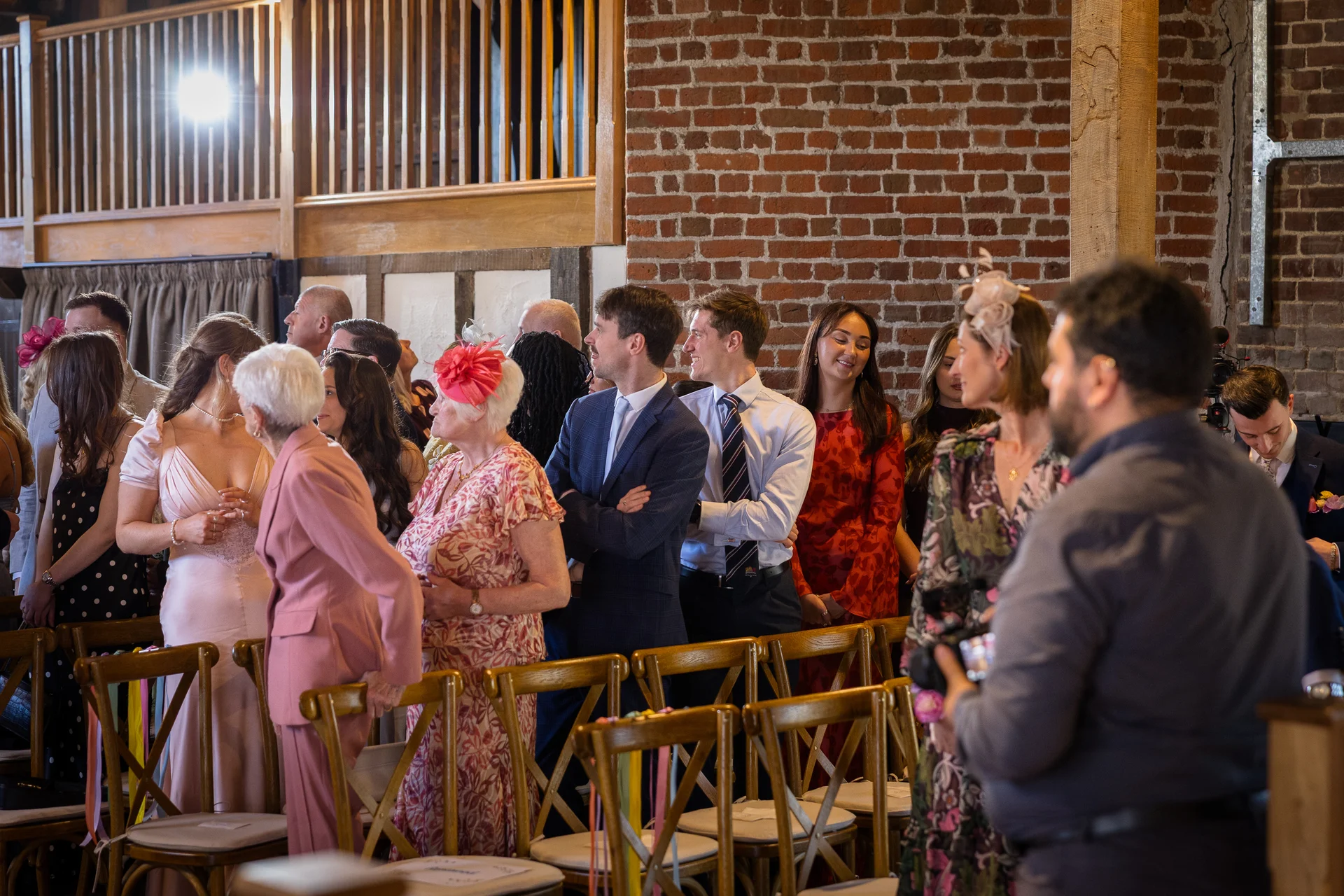 Wedding guests dressed in smart attire and fascinators mingle inside a rustic barn venue featuring exposed brick walls and wooden chairs.