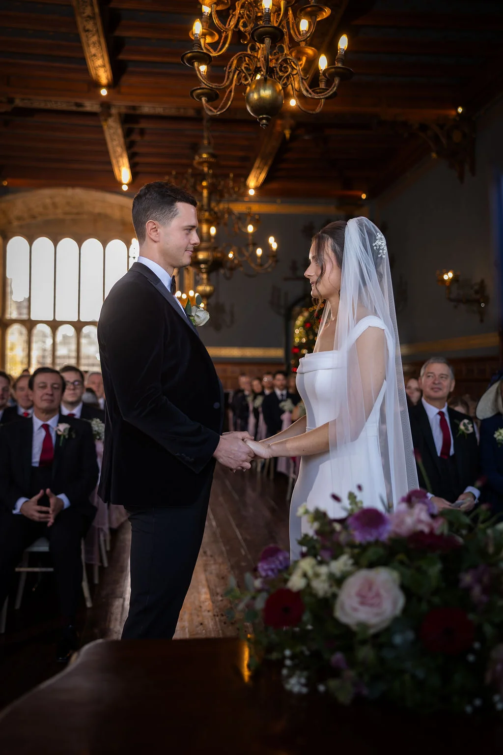 A bride and groom lovingly hold hands during their candlelit indoor ceremony inside a historic manor house featuring wooden beamed ceilings and traditional chandeliers.
