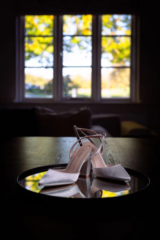 A pair of satin bridal block heel shoes resting on a round mirror surface in front of a bright window.