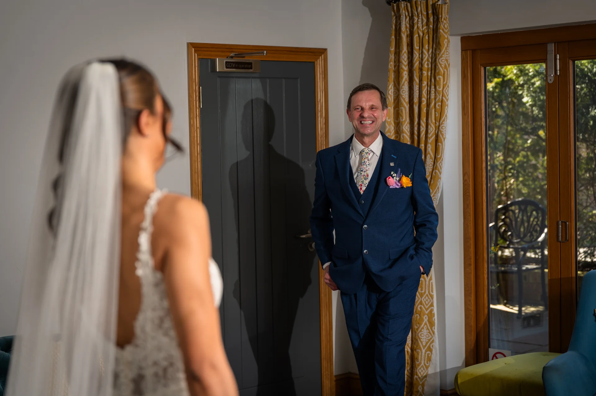 A smiling father in a navy blue suit looks proudly at his daughter, who stands in her wedding dress with a veil, during a first look moment.